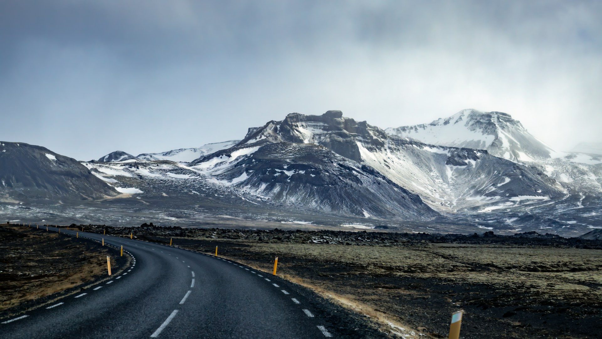 A car on Iceland's Ring Road curving along a black-sand coast beneath a waterfall and low cloud, with a single-lane bridge ahead